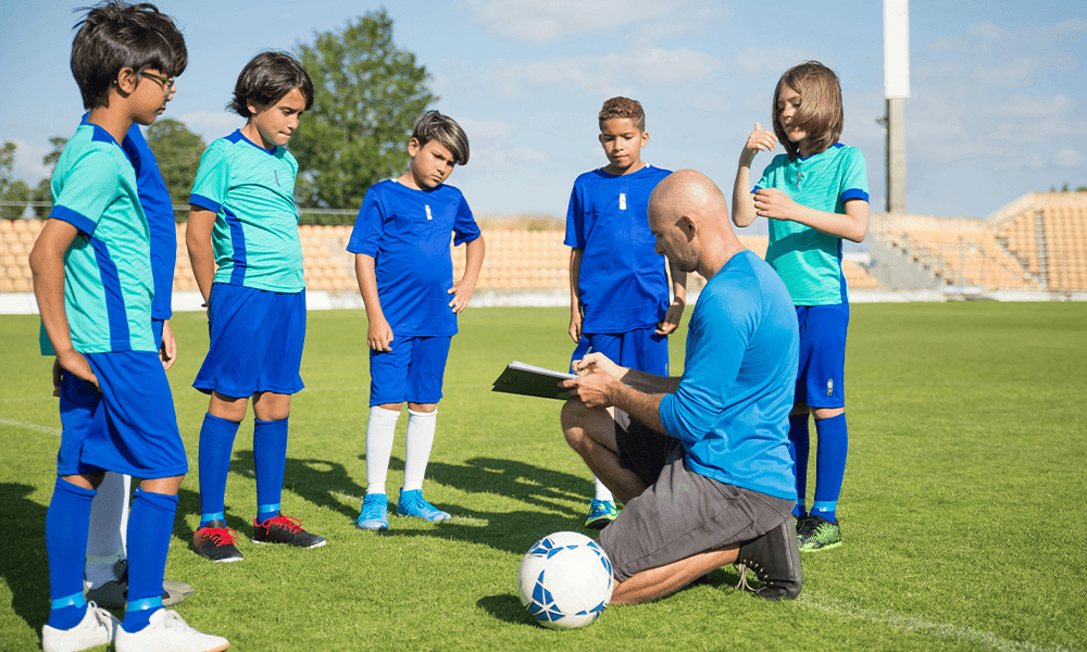 A coach in a blue uniform gives instructions to young soccer players on a sunny field. The players, also in blue uniforms, attentively listen.