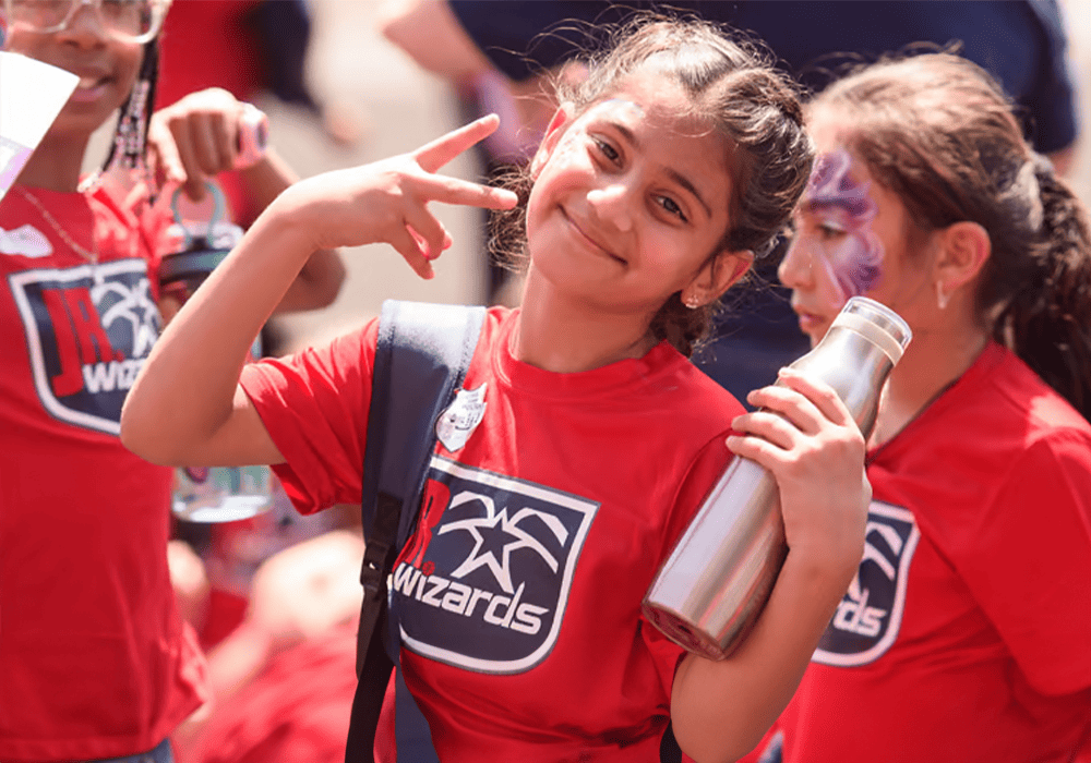 A group of children wearing Washington Wizards t-shirts, one child in the foreground smiling and gesturing a peace sign, during a sunny outdoor event.