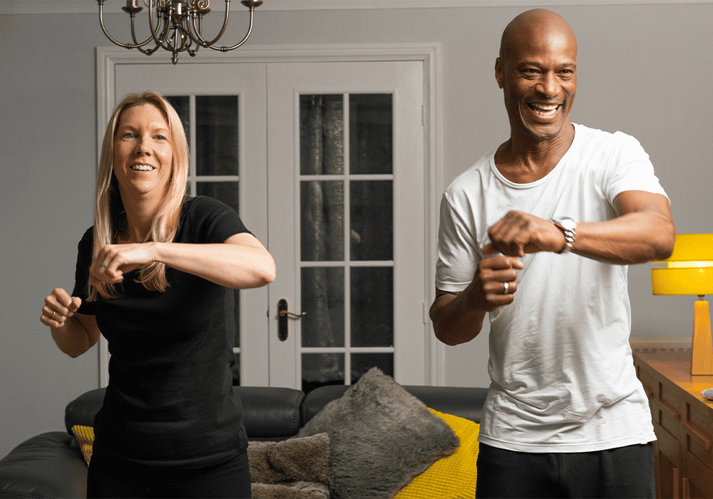 Two adults smiling and exercising in a home setting.