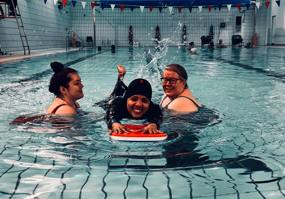 Three individuals are enjoying time in an indoor swimming pool, one of them playfully splashing water with a kickboard. They appear to be smiling and having fun together. Flags hang in the background.